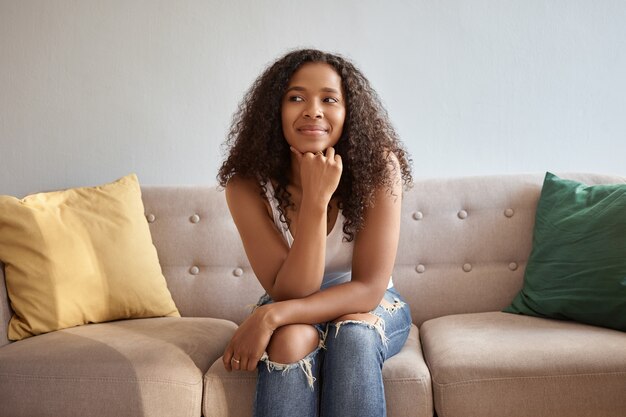 Charming young Afro American woman with loose curly hair sitting on couch at home wearing trendy ripped jeans and white tank top, looking away with pensive expression, thinking about plans on weekend