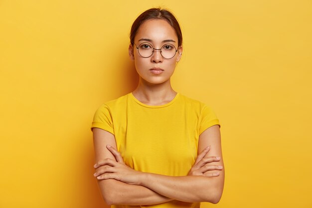 Charming young Asian woman has fresh skin, looks  confidently, has serious expression, keeps hands crossed over chest, wears optical glasses and yellow t shirt, being deep in thoughts