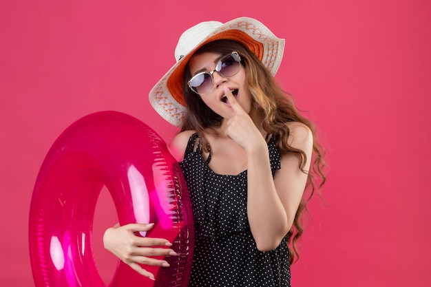 핑크 공간 위에 서있는 입술에 손가락으로 놀란 풍선 반지를 들고 선글라스를 착용하는 여름 모자에 폴카 도트 드레스에 매력적인 젊은 아름다운 여행자 소녀