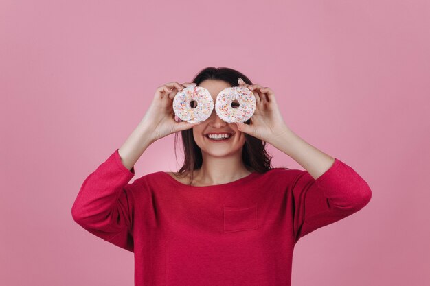 Очаровательная молодая брюнетка держит розовые пончики перед ее глазами, позирует в розовой комнате