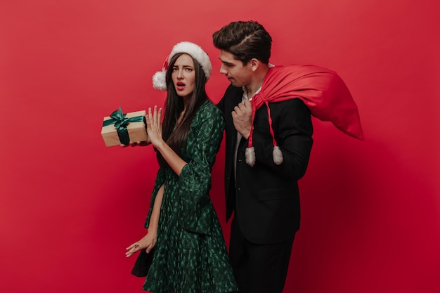 Charming young brunette with green dress and Santa hat declining gift box from her boyfriend in black suit Pair posing isolated on red background