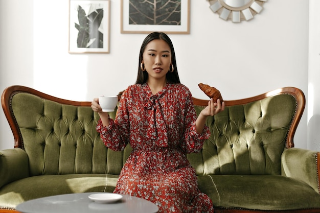 Charming young brunette woman in red floral dress looks into camerasits on soft velvet green sofa holds white cup of tea and croissant