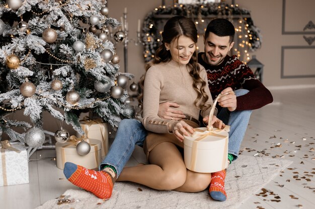 Charming young couple in cozy home clothes opens present boxes before a Christmas tree