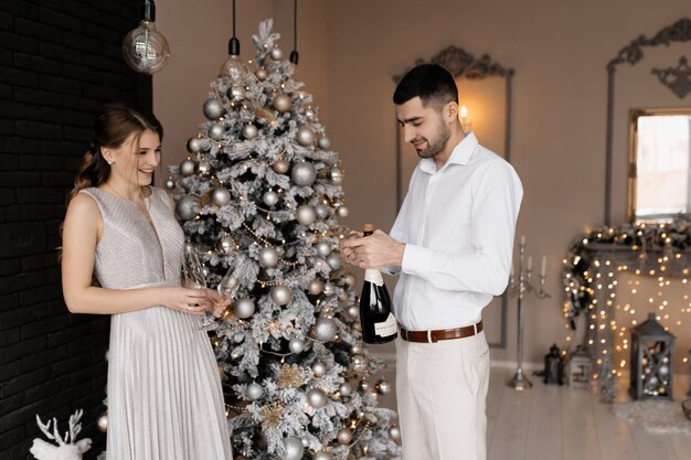 Очаровательная молодая пара в модной одежде позирует с бокалами для шампанского перед елкой