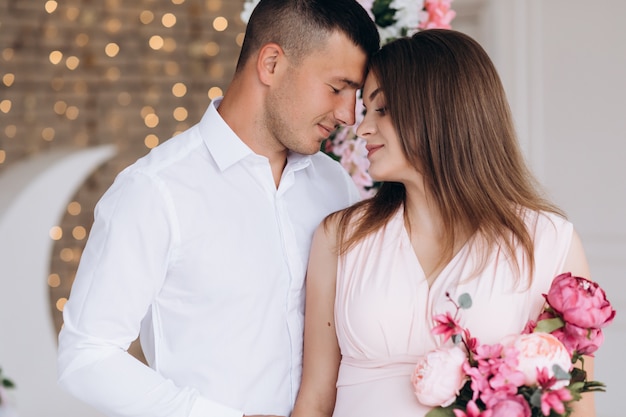 Charming young expecting couple poses in a white studio rich decorated with pink flowers