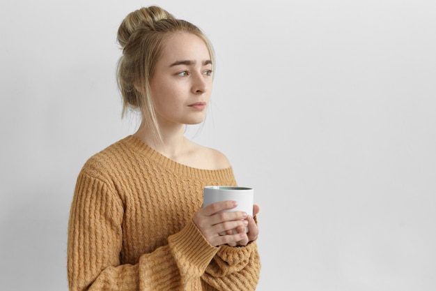 Affascinante giovane donna che indossa un'acconciatura disordinata e un pullover lavorato a maglia oversize in posa sul muro grigio bianco, tenendo in mano una grande tazza, bevendo tè, caffè, cioccolata calda o cioccolata calda al mattino