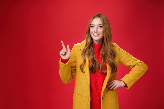 Charming young female in yellow coat holding hand on waist and pointing at upper left corner, smiling broadly as showing or promoting interesting proposal over red background, in good mood.