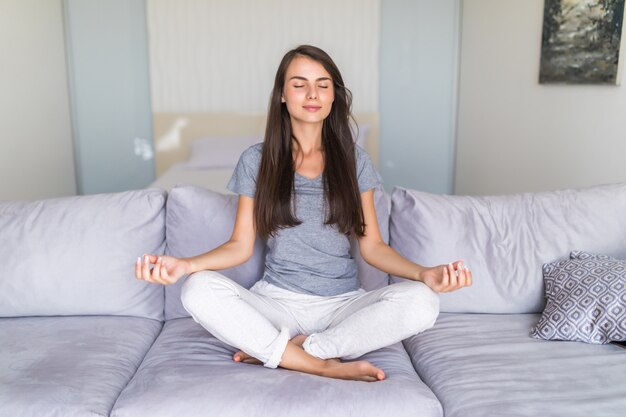 Charming young lady in casual clothes meditating couch cross-legged, doing yoga exercises in living room in morning, relaxing and making autosuggestion practices to get positive thinking