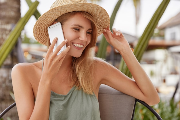 Charming young lovely woman has telephone conversation with boyfriend via smart phone, sits alone in coffee shop, wears casual clothing and summer hat, has tanned healthy skin and shining smile