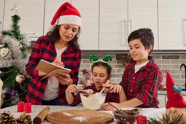 サンタ​の​帽子​を​かぶった​魅力的​な​若い​お母さん​は​、​子供たち​が​ボウル​に​材料​を​混ぜている​間​、​彼女​の​子供たち​に​レシピ​を​指示します​。​愛情​深い​母親​と​甘い​子供たち​-​男の子​と​女の子​が​家​で​一緒​に​料理する​クリスマス​クッキー