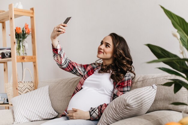 格子縞のシャツと白いTシャツの魅力的な若い妊婦は自分撮りを取り、電話を保持します