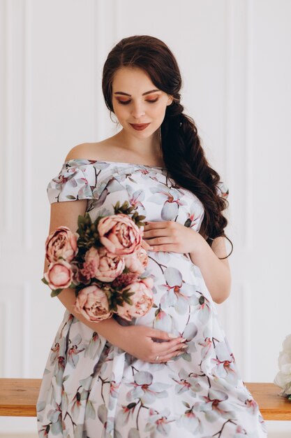 Charming young pregnant woman poses in a studio with pink flowers
