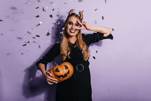 Очаровательная молодая женщина в черном платье, наслаждаясь карнавалом хеллоуина. фотография улыбающейся девушки-вампира, держащей оранжевую тыкву.