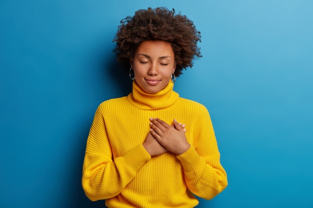 Charming young woman keeps hands on chest to express gratitude, makes heart sign, expresses beneficence, feels thankful, keeps eyes closed