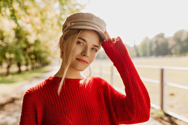 빨간 스웨터와 공원에서 좋은 찾고 가벼운 모자에 매력적인 젊은 여자. 가을에 멋진 액세서리와 트렌디 한 옷을 입고 아름다운 금발.
