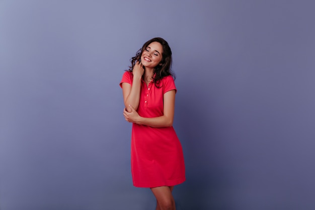 Очаровательная молодая женщина улыбается с закрытыми глазами на фиолетовой стене. Фотография беспечной кудрявой девушки в модном красном платье в помещении.