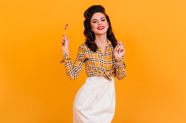 Charming young woman in vintage attire holding candy. Smiling pinup girl with lollipop standing on yellow background.