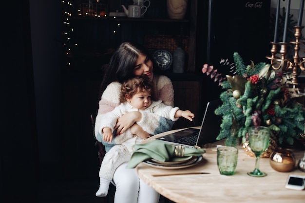 Charming young woman with cute kid sitting at table and looking at laptop screen