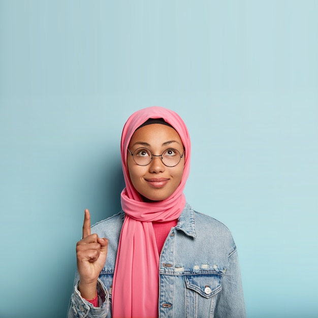 Charming young woman with dark healthy skin, looks and points up, indicates with index finger, wears pink hijab, fashionable jacket, stands over blue wall, advertises copy space, shows right way