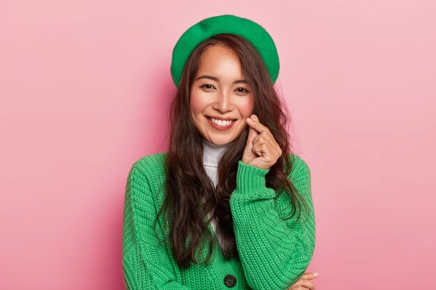 Charming young woman with dark long hair makes korean love sign, shapes heart with fingers, wears bright fashionable green beret and jumper on buttons