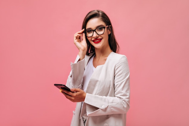 Charming young woman with red lips in beige outfit and eyeglasses looks into camera and holds smartphone on isolated pink background.