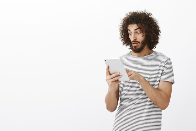Chart of company raised unexpectedly. Shocked and pleased handsome male student with curly hair and beard, dropping jaw while scrolling news feed in digital tablet