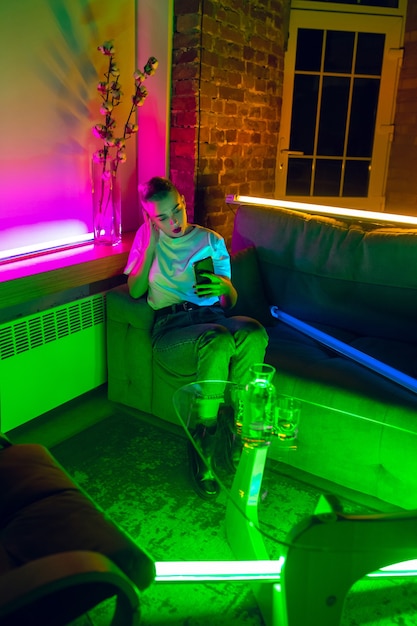 Chatting. Cinematic portrait of stylish woman in neon lighted interior. Toned like cinema effects, bright neoned colors. Caucasian model using smartphone in colorful lights indoors. Youth culture.