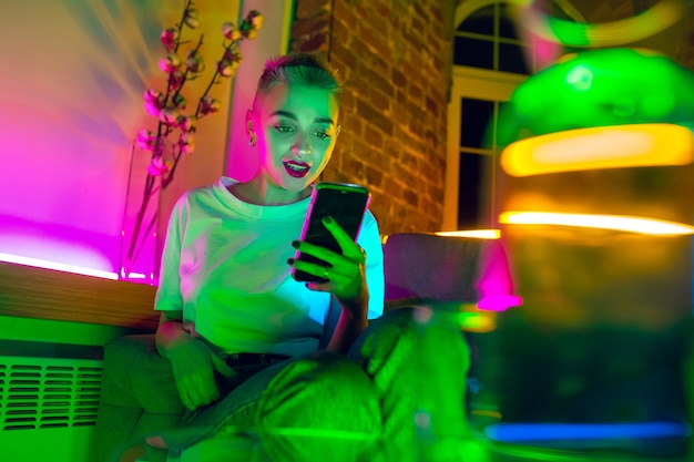 Chatting. Cinematic portrait of stylish woman in neon lighted interior. Toned like cinema effects, bright neoned colors. Caucasian model using smartphone in colorful lights indoors. Youth culture.