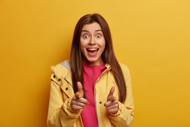 Check it out. Positive joyful young pretty European woman points both index fingers , says yeah, dressed in casual anorak, picks you, isolated on yellow wall, has optimistic gaze