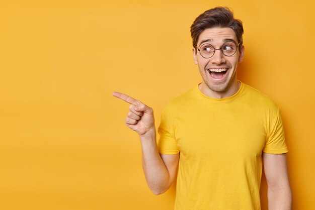 Check this out Cheerful handsome young man points index finger away demonstrates advertisement wears transparent round spectacles and t shirt isolated over yellow background Promotion concept