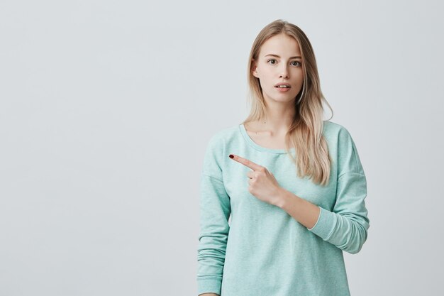 Check this out Cropped attractive good-looking excited young woman in blue sweater pointing fingers aside having surprised look, with cheerful and excited expression of face.