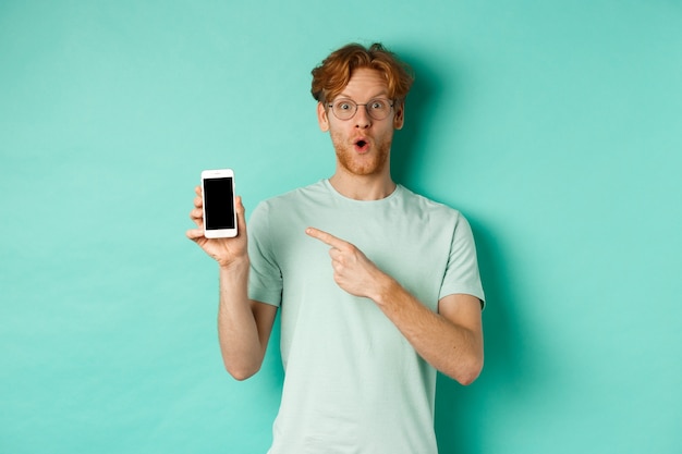 これをチェックしてください。空白のスマートフォンの画面に指を指して、オンラインプロモーションを表示し、ターコイズブルーの背景に驚いて立っている眼鏡のハンサムな赤毛の男