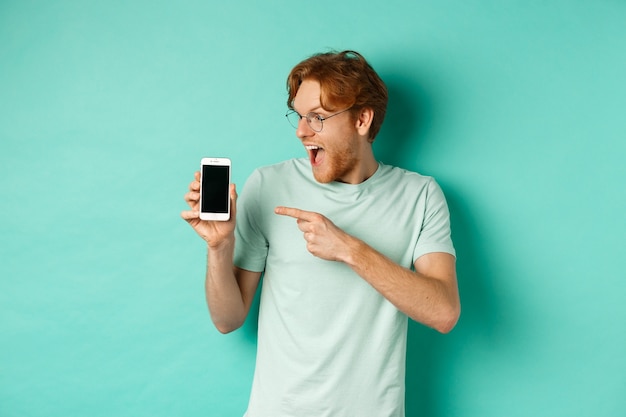 これをチェックしてください。空白のスマートフォンの画面に指を指して、オンラインプロモーションを表示し、ターコイズブルーの背景に驚いて立っている眼鏡のハンサムな赤毛の男