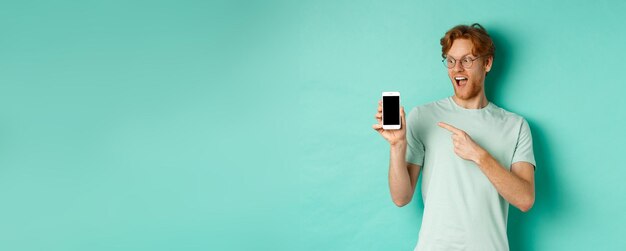 Foto gratuita dai un'occhiata a questo bel ragazzo dai capelli rossi con gli occhiali che punta il dito sullo schermo vuoto dello smartphone in mostra