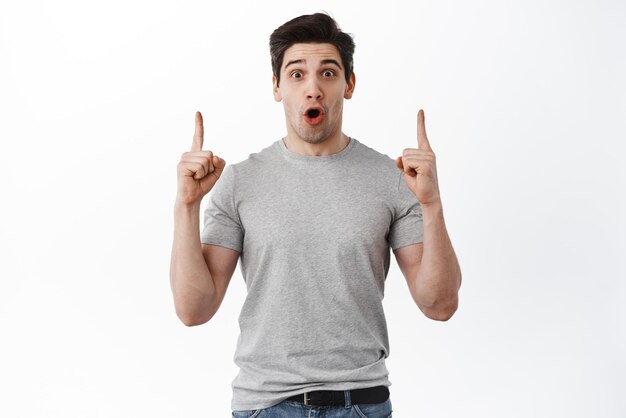 Check this out Impressed 30s man say wow pointing fingers up at advertisement stare amazed at camera showing promo banner standing over white background