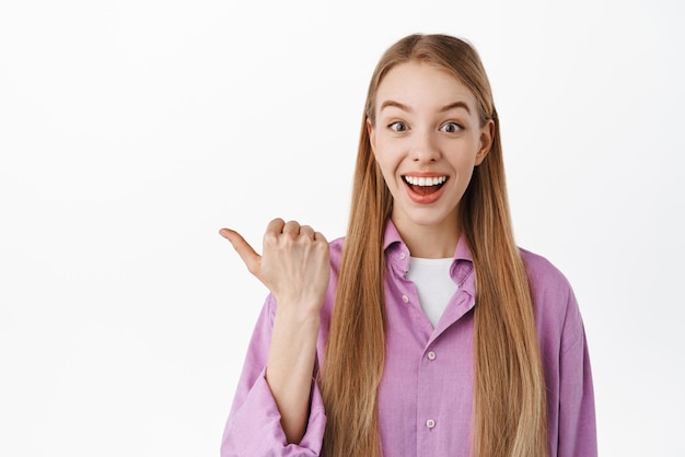 Check this out Smiling happy woman with long blond hair pointing thumb left advertising special discounts showing promo deal standing against white background