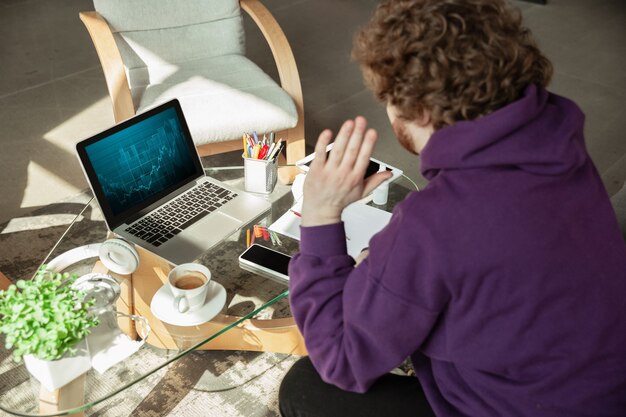 Проверка графиков. Человек, работающий из дома во время карантина коронавируса или COVID-19, концепция удаленного офиса. Молодой бизнесмен, менеджер, выполняющий задачи со смартфоном, ноутбуком, планшетом, проводит онлайн-конференцию.