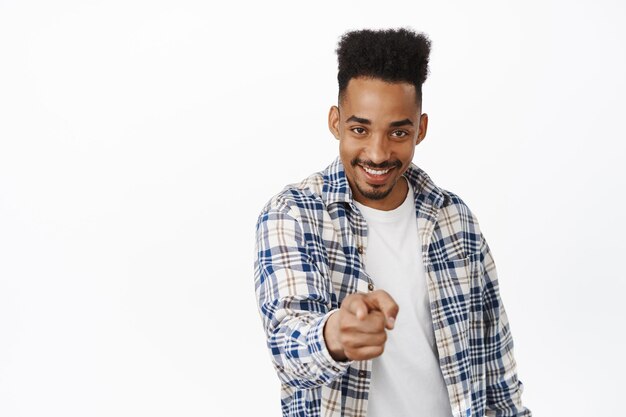 Cheeky african american man smiling, pointing at camera with confidence smug face, choosing you, inviting or recruiting to job, beckon join him, standing over white background, picking.