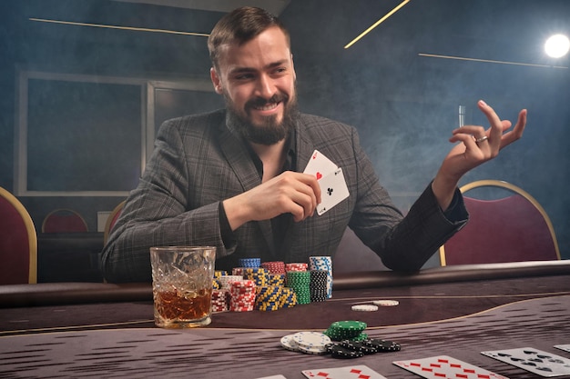 cheeky brunette man in a classic suit is playing poker sitting at the table at casino in a smoke. he is making bets waiting for a big win. gambling for money. games of fortune.