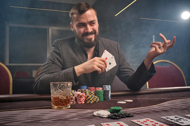 고전적인 양복을 입은 건방진 갈색 머리 남자가 연기 속에서 카지노 테이블에 앉아 포커를 하고 있습니다. 그는 큰 승리를 기다리며 내기를 하고 있습니다. 돈을 위한 도박. 행운의 게임.