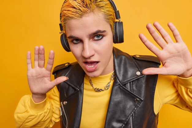Cheeky confident teenage girl has bright makeup trendy hairstyle keeps palms forward camera raised listens music in black wireless headphones wears fashionable outfit isolated on yellow wall