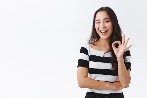 Cheeky feminine asian girlfriend keep everything under control, standing relaxed and chill with no worries, showing okay, confirmation or approval gesture, wink hinting and smiling, white background