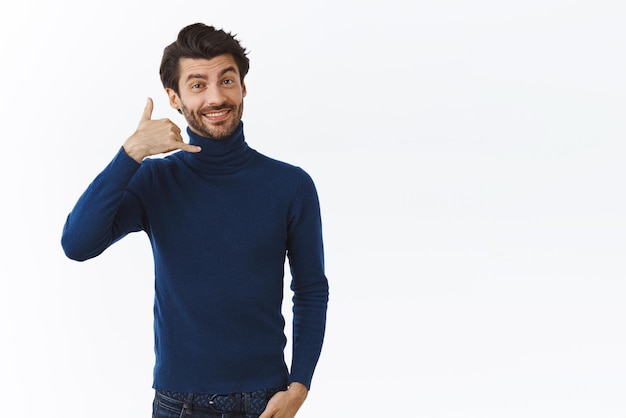 Cheeky flirty and coquettish handsome stylish bearded man in high neck sweater smiling sassy making phone gesture near ear asking cute women give him call asking for number white background