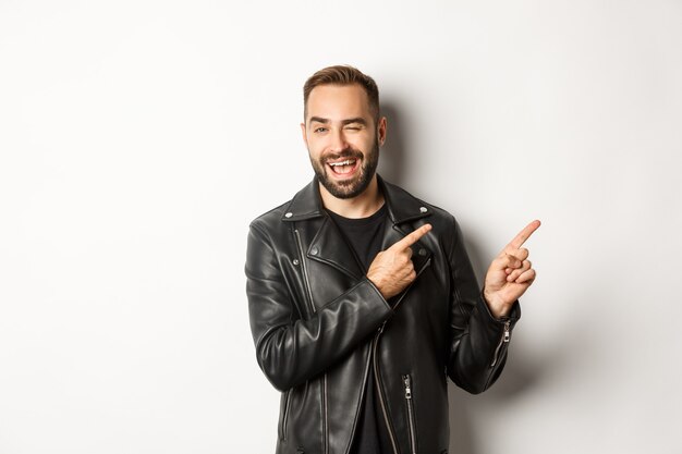 Cheeky guy in leather biker jacket, winking flirty and pointing at upper right corner copy space, white background.