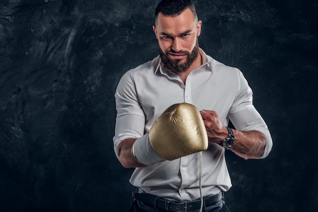 Cheeky handsome man in white shirt and with golden boxing glove is standing at dark studio.