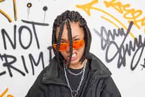 Free photo cheeky hipster girl looks with confident impudent expression at camera feels cool belongs to youth subculture wears trendy orange sunglasses and jacket poses against graffiti wall