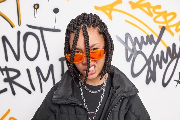 Cheeky hipster girl looks with confident impudent expression at camera feels cool belongs to youth subculture wears trendy orange sunglasses and jacket poses against graffiti wall