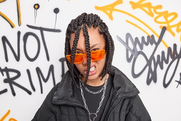 Cheeky hipster girl looks with confident impudent expression at camera feels cool belongs to youth subculture wears trendy orange sunglasses and jacket poses against graffiti wall