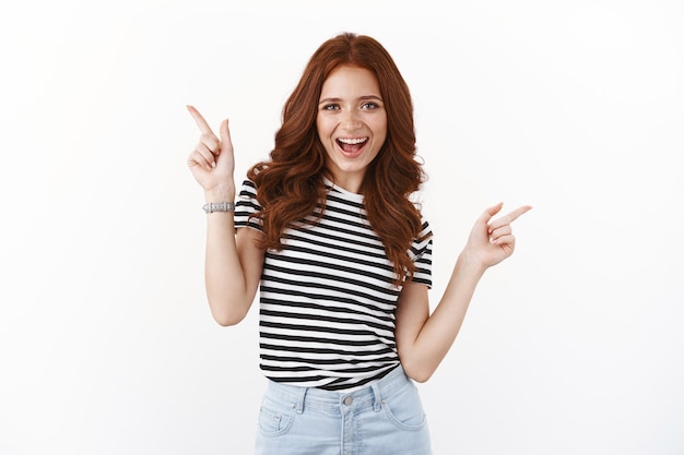 Cheeky playful redhead girl dancing upbeat, pointing sideways, showing left right copy space promos, smiling joyfully, invite try offer, recommend good spots place advertisement, white wall