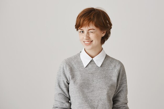 Cheeky smiling redhead girl with short haircut posing against the white wall